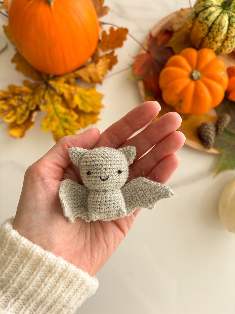 Op de afbeelding: Een kleine, lichtgrijze gehaakte vleermuis knuffel met puntige oren, uitgestrekte vleugels en een geborduurd gezicht met twee zwarte kraaloogjes, een eenvoudig gebogen mondje en roze blosjes op de wangen, gemaakt van gestructureerd garen.