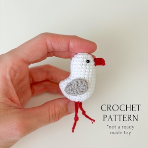 Puede incluir: Un pájaro blanco tejido a crochet con pico y patas rojas. El pájaro tiene un detalle de ala gris. Patrón de crochet, no un juguete listo para usar.