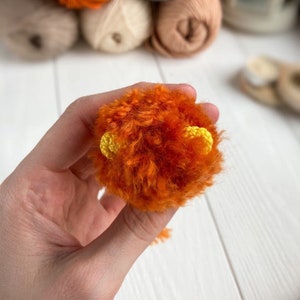K&ouml;nnte beinhalten: Ein kleiner orangefarbener Pompon mit gelben Garn-Ohren. Der Pompon wird in einer Hand gehalten.