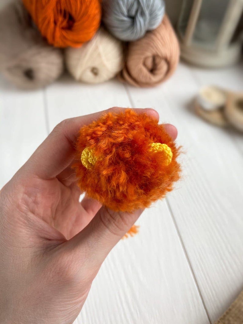 K&ouml;nnte beinhalten: Ein kleiner orangefarbener Pompon mit gelben Garn-Ohren. Der Pompon wird in einer Hand gehalten.