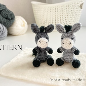 May include: Two gray crocheted donkey amigurumi toys with black eyes and hooves. The donkeys are sitting on a white fluffy surface. The text "PATTERN" is visible in the image. The text "not a ready made item" is also visible in the image.