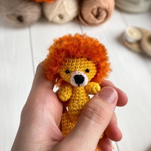 K&ouml;nnte beinhalten: Ein kleiner geh&auml;kelter L&ouml;we mit orangefarbenem Fell, einem gelben K&ouml;rper und schwarzen Augen. Der L&ouml;we wird in einer Hand gehalten.