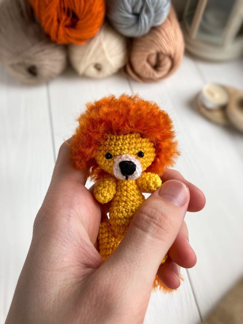 K&ouml;nnte beinhalten: Ein kleiner geh&auml;kelter L&ouml;we mit orangefarbenem Fell, einem gelben K&ouml;rper und schwarzen Augen. Der L&ouml;we wird in einer Hand gehalten.