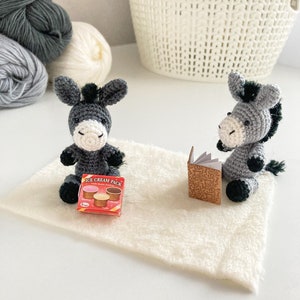 May include: Two gray crocheted donkey toys sitting on a white fuzzy rug. One donkey is holding a small book and the other is holding a small box of ice cream.