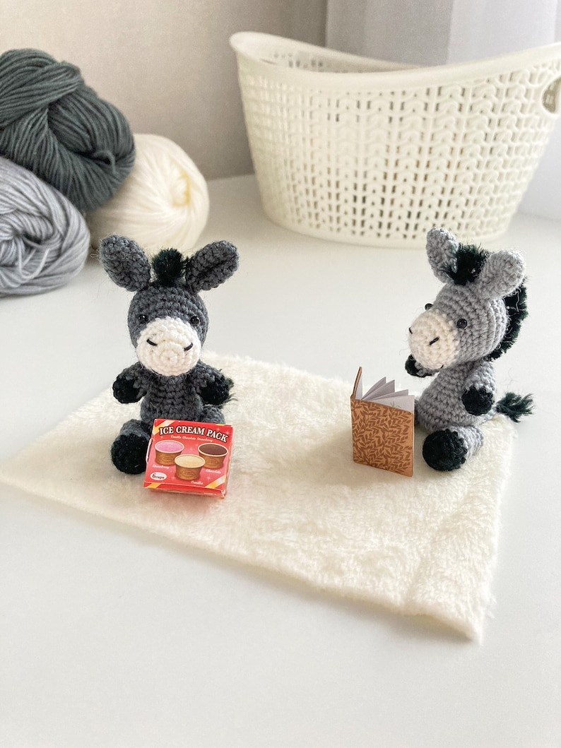 May include: Two gray crocheted donkey toys sitting on a white fuzzy rug. One donkey is holding a small book and the other is holding a small box of ice cream.