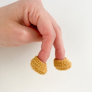 May include: Two crocheted fingertip covers in a mustard yellow color. The covers are worn on two fingers of a hand.