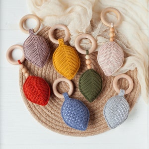 May include: A set of seven crocheted leaf-shaped teethers with wooden rings and beads. The teethers are in various colors, including red, orange, yellow, green, blue, and gray.
