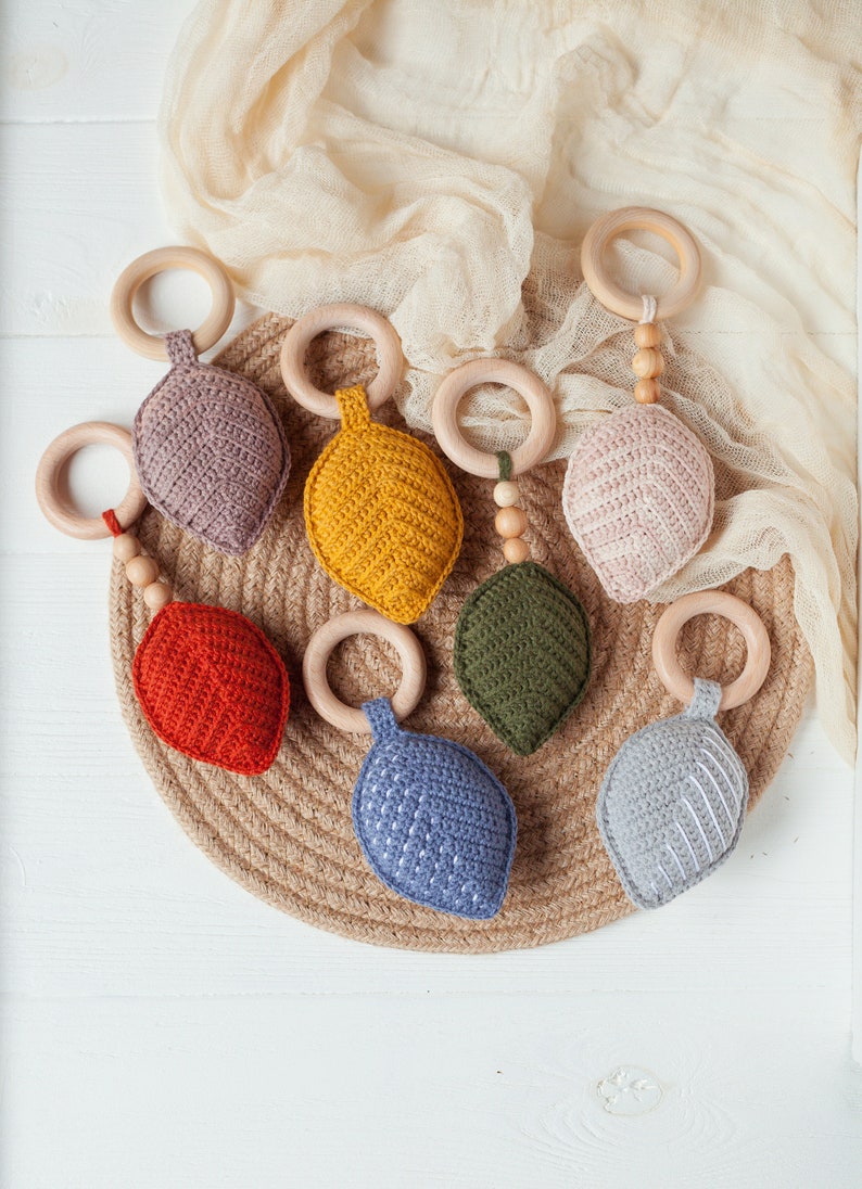 May include: A set of seven crocheted leaf-shaped teethers with wooden rings and beads. The teethers are in various colors, including red, orange, yellow, green, blue, and gray.
