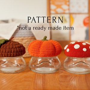 May include: Three crocheted jar lids in the shapes of a pumpkin, mushroom, and acorn. The lids are orange, red, and brown. The lids are sitting on top of glass jars. The text "PATTERN *not a ready made item*" is displayed above the jars.