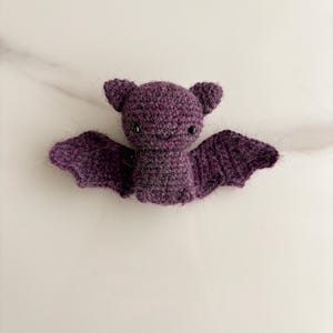 Op de afbeelding: Een kleine, donkerpaarse gehaakte vleermuis knuffel met een pluizige textuur. De vleermuis heeft twee puntige oren, twee kleine zwarte kraaloogjes, een geborduurde glimlach en twee uitgestrekte vleugels van garen.