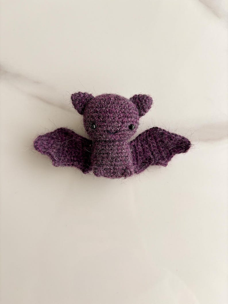 Op de afbeelding: Een kleine, donkerpaarse gehaakte vleermuis knuffel met een pluizige textuur. De vleermuis heeft twee puntige oren, twee kleine zwarte kraaloogjes, een geborduurde glimlach en twee uitgestrekte vleugels van garen.