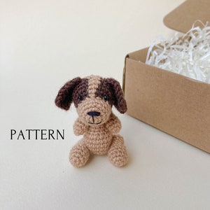 Peut inclure: Un petit jouet en crochet en forme de chien, de couleur marron et beige, avec des yeux noirs. Le jouet est posé sur une surface blanche. Le mot "PATTERN" est visible dans le coin inférieur gauche de l'image.