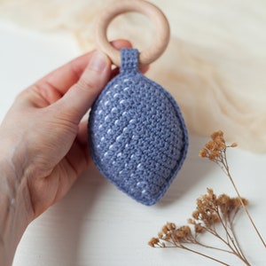 May include: A blue crocheted baby teether with a white wooden ring. The teether is shaped like a leaf and has a textured surface.