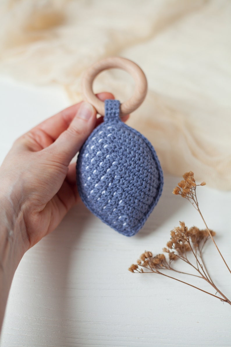 May include: A blue crocheted baby teether with a white wooden ring. The teether is shaped like a leaf and has a textured surface.