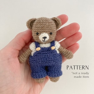 May include: Crocheted brown teddy bear wearing blue overalls with white shirt and wooden buttons.  The bear is holding a small white flower.  PATTERN *not a ready made item*