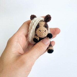 May include: A small, handmade crochet horse figurine. The horse features a brown and tan body with a white mane and tail. The horse is held in a hand against a white background. The horse is a miniature version of a real horse.