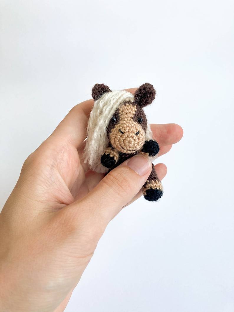 May include: A small, handmade crochet horse figurine. The horse features a brown and tan body with a white mane and tail. The horse is held in a hand against a white background. The horse is a miniature version of a real horse.