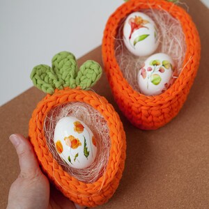 May include: Two crocheted orange carrot-shaped baskets with green tops, each holding white Easter eggs decorated with floral designs.