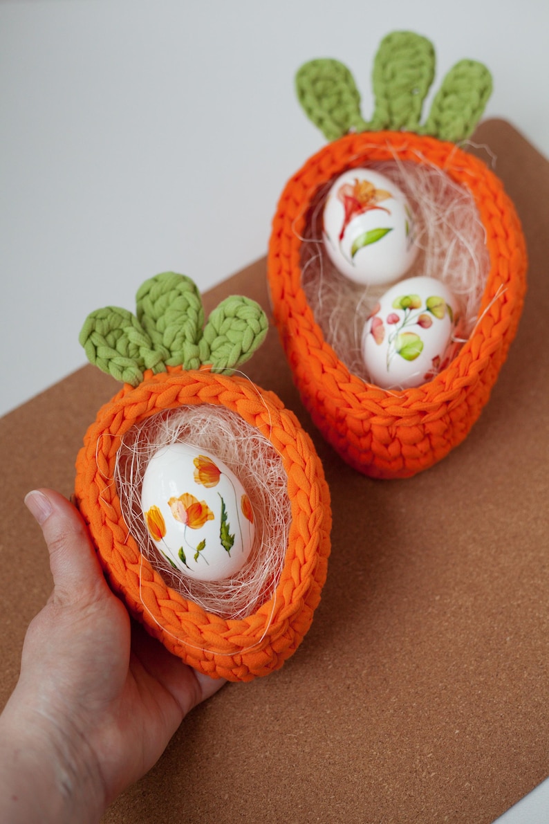 May include: Two crocheted orange carrot-shaped baskets with green tops, each holding white Easter eggs decorated with floral designs.