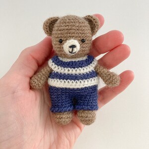 May include: A small brown crocheted teddy bear wearing a blue and white striped shirt and blue trousers.