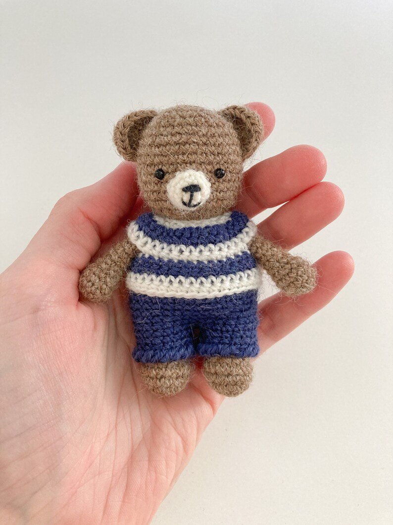 May include: A small brown crocheted teddy bear wearing a blue and white striped shirt and blue trousers.