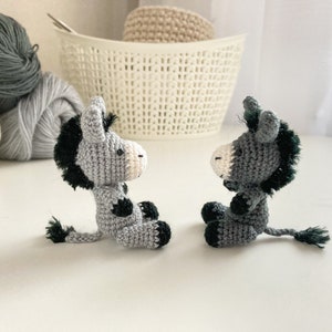 May include: Two gray crocheted donkey figurines with black accents and black yarn tails. The donkeys are facing each other.