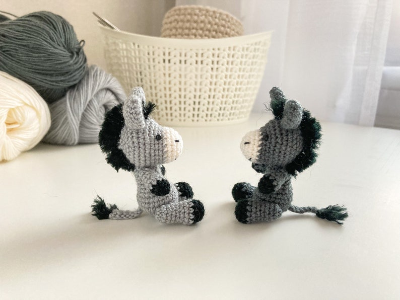 May include: Two gray crocheted donkey figurines with black accents and black yarn tails. The donkeys are facing each other.