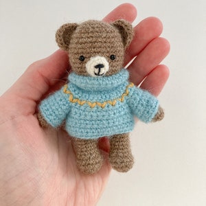 May include: A small brown crocheted teddy bear wearing a blue jumper with a yellow trim. The bear has black eyes and a white nose.
