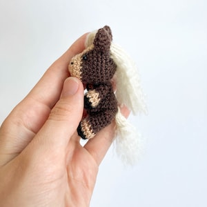 May include: A small, handmade crochet horse figurine. The horse is primarily brown with tan and black accents, and a white mane and tail. The miniature horse is held in a hand against a white background. The horse is a cute, whimsical toy.