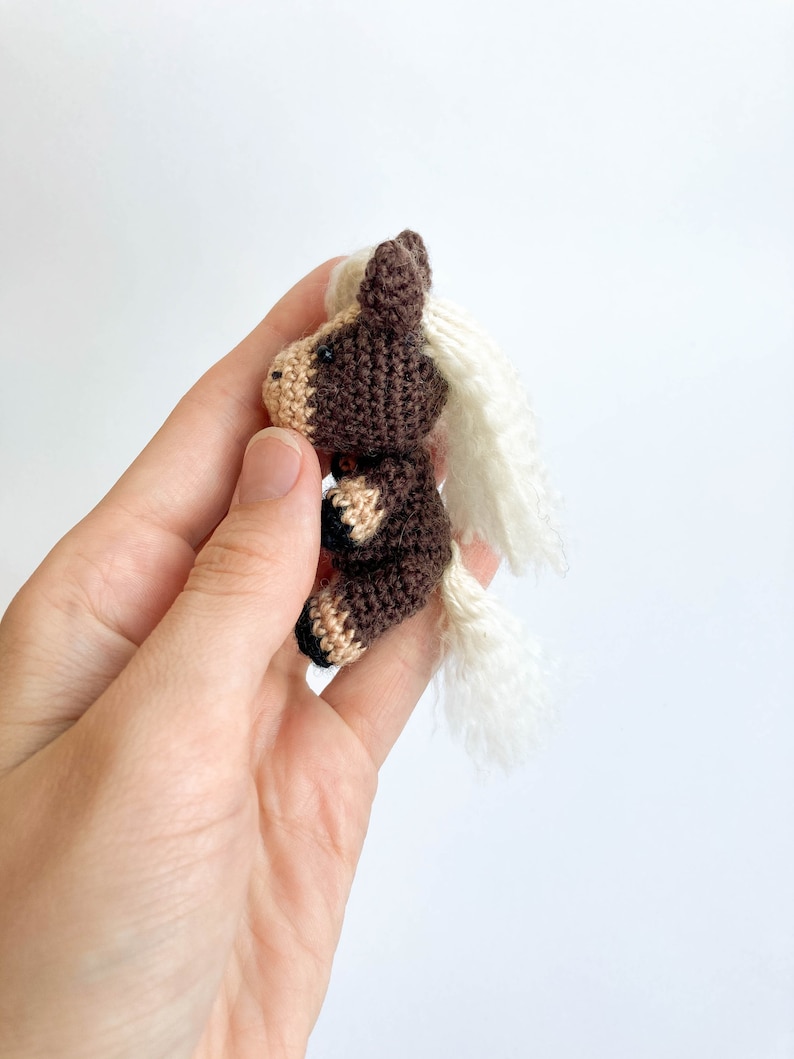 May include: A small, handmade crochet horse figurine. The horse is primarily brown with tan and black accents, and a white mane and tail. The miniature horse is held in a hand against a white background. The horse is a cute, whimsical toy.
