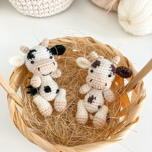 May include: Two crocheted cow plushies with black and brown spots, sitting in a woven basket with straw-like filling.