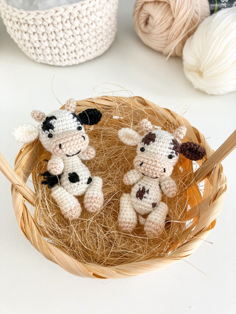 May include: Two crocheted cow plushies with black and brown spots, sitting in a woven basket with straw-like filling.