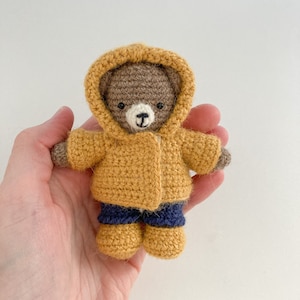 May include: A small crocheted brown bear wearing a yellow raincoat and blue pants. The bear is being held in a person's hand.