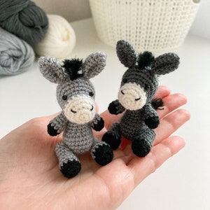 May include: Two small gray crocheted donkey figurines with black hooves and eyes. The donkeys are sitting on a hand.