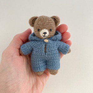 May include: A small brown crocheted teddy bear wearing a blue crocheted hooded jumpsuit.