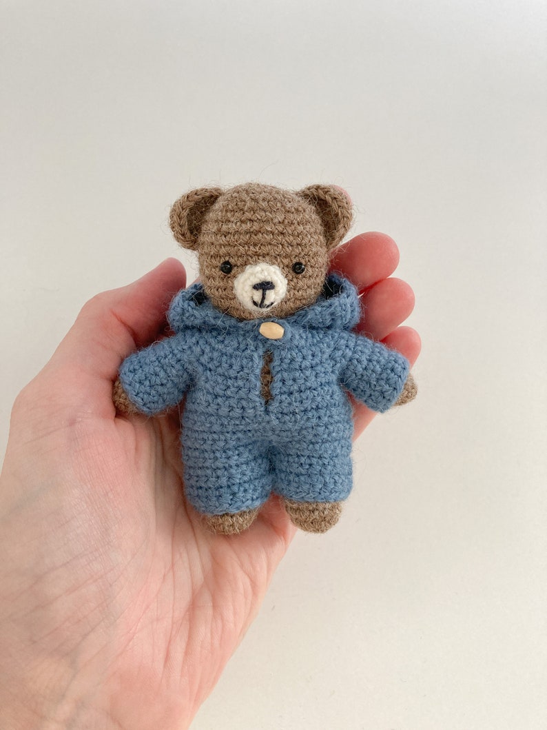 May include: A small brown crocheted teddy bear wearing a blue crocheted hooded jumpsuit.
