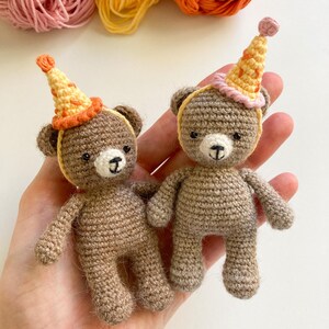 May include: Two small brown crocheted teddy bears wearing party hats. One hat is pink and yellow, the other is orange and yellow.