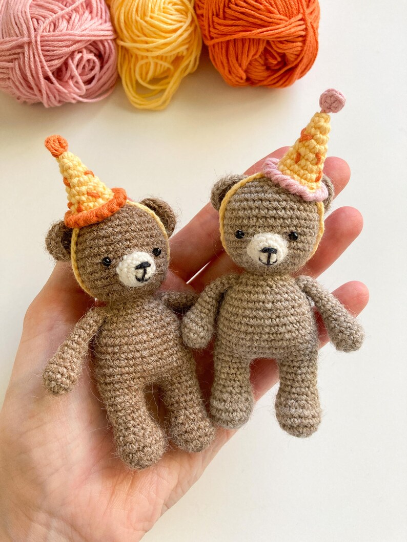 May include: Two small brown crocheted teddy bears wearing party hats. One hat is pink and yellow, the other is orange and yellow.
