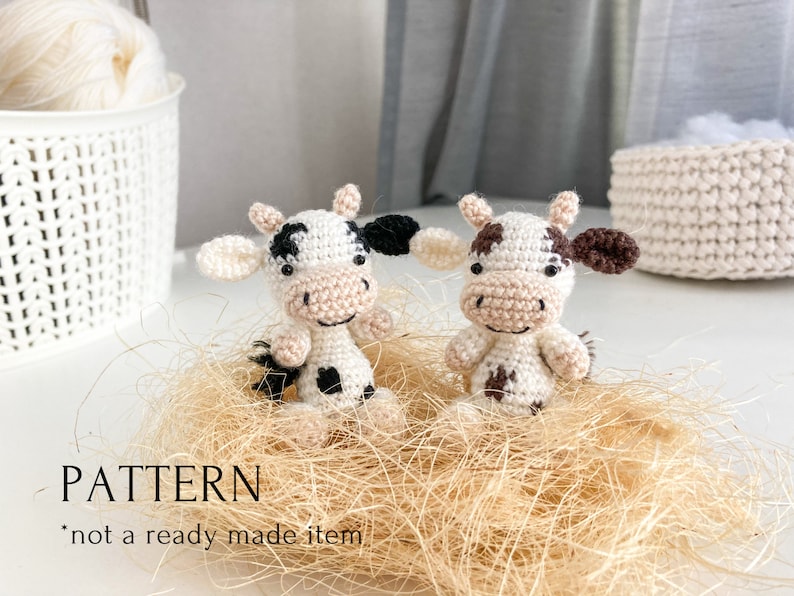 May include: Two crocheted cow amigurumi toys with black and white spots and brown ears. The toys are sitting in a nest of tan straw. The text "PATTERN *not a ready made item*" is at the bottom of the image.