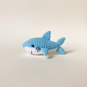 May include: A light blue crocheted shark toy with white detailing around the mouth. The shark has black eyes and a stitched smile.