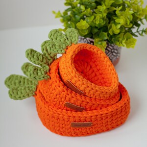 May include: Three orange crocheted baskets in the shape of carrots, with green crocheted tops. The baskets are stacked on top of each other, with the smallest basket on top. The baskets have brown leather tags that say "Hand Made".