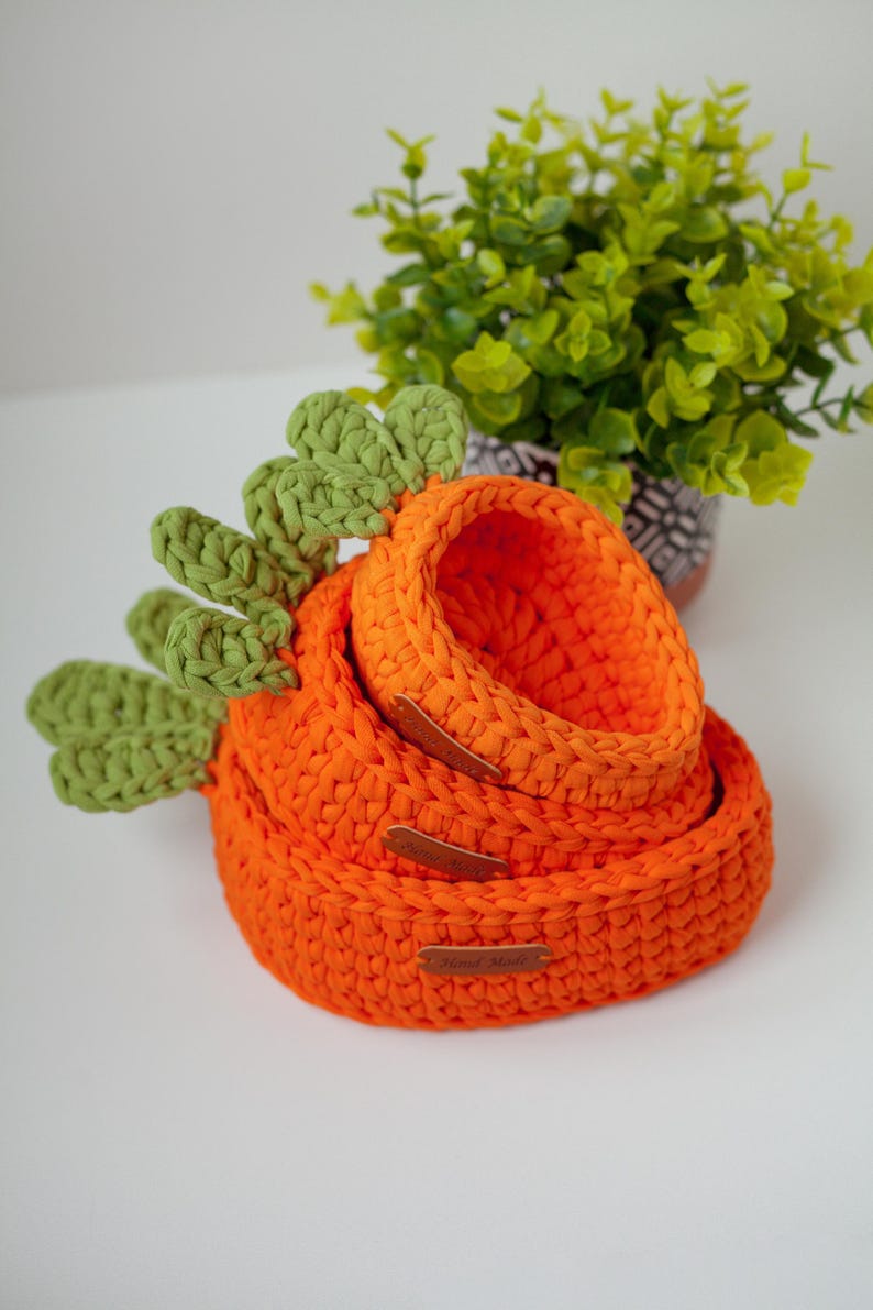May include: Three orange crocheted baskets in the shape of carrots, with green crocheted tops. The baskets are stacked on top of each other, with the smallest basket on top. The baskets have brown leather tags that say "Hand Made".