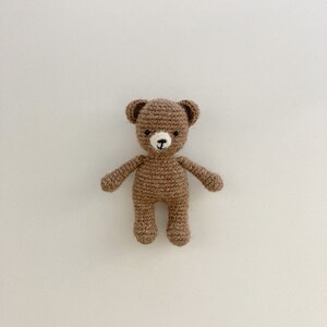 K&ouml;nnte beinhalten: Ein brauner geh&auml;kelter Teddyb&auml;r mit wei&szlig;en Akzenten f&uuml;r Nase und Augen. Der B&auml;r steht auf einem wei&szlig;en Hintergrund.