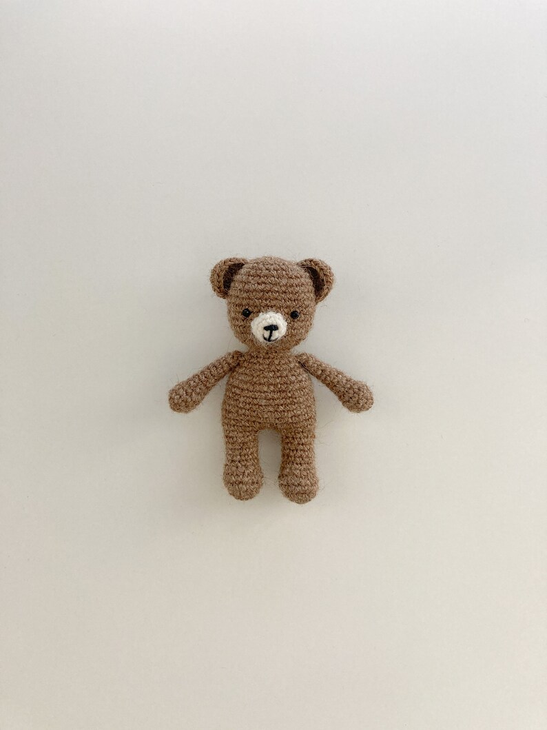 K&ouml;nnte beinhalten: Ein brauner geh&auml;kelter Teddyb&auml;r mit wei&szlig;en Akzenten f&uuml;r Nase und Augen. Der B&auml;r steht auf einem wei&szlig;en Hintergrund.
