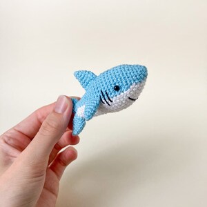 May include: A small crocheted blue shark toy with white accents and black eyes. The shark is being held in a hand.