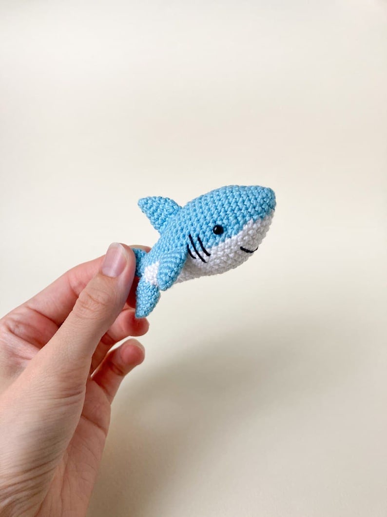 May include: A small crocheted blue shark toy with white accents and black eyes. The shark is being held in a hand.