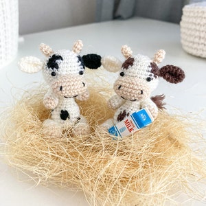 May include: Two crocheted white cow figurines with black spots, one holding a miniature carton of milk that says "Fresh Milk" on it. The cows are sitting in a nest of tan straw.
