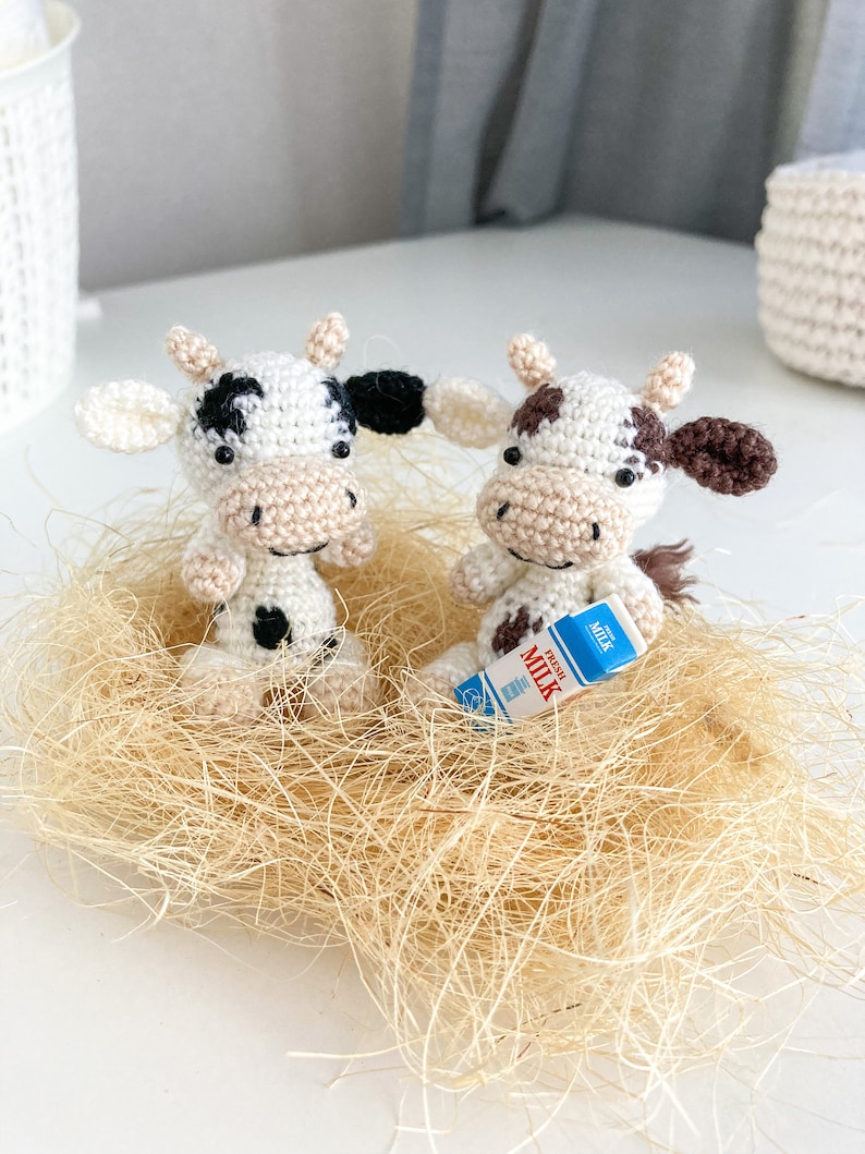 May include: Two crocheted white cow figurines with black spots, one holding a miniature carton of milk that says "Fresh Milk" on it. The cows are sitting in a nest of tan straw.