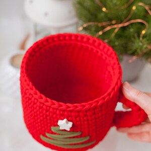 Pu&ograve; includere: Tazza in crochet rosso con un design a forma di albero di Natale verde e una stella bianca in cima.