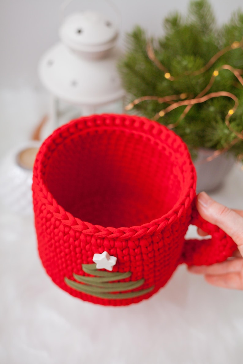 Pu&ograve; includere: Tazza in crochet rosso con un design a forma di albero di Natale verde e una stella bianca in cima.